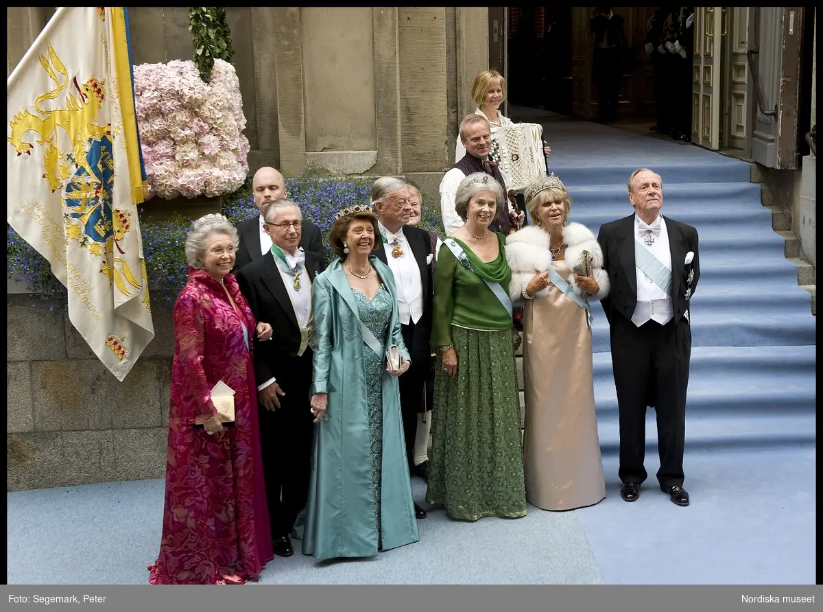 Kronprinsessbröllopet. Dokumentation av bröllopet mellan Kronprinsessan Victoria och herr Daniel Westling 19 juni 2010. Gästerna anländer till Storkyrkan. Från vänster; Prinsessan Birgittas make, Johan Georg av Sigmaringen i frack och ordenstecken. Prinsessan Birgitta i champagnefärgad sidenklänning med vit pälsjacka, drottning Sofias diadem, smycken, kuvertväska och ordenstecken. Prinsessan Margaretha i grön klänning med brokad och transparent blus, diadem med akvamariner, smycken, kuvertväska och ordenstecken. Prinsessan Désirées make Nicklas Silfverschiöld i frack. Prinsessan Désirée i turkos klänning med brokad och aftoncape i siden i samma nyans, juvelgarnityr med ametister och briljanter, vit kuvertväska och ordenstecken. Prinsessan Christinas make, Tord Magnuson i frack. Prinsessan Christina i cyklamenröd klänning i blank sidensatin med brokadvävd aftonkappa, Karl Johan-diademet, smycken, kuvertväska och ordenstecken.