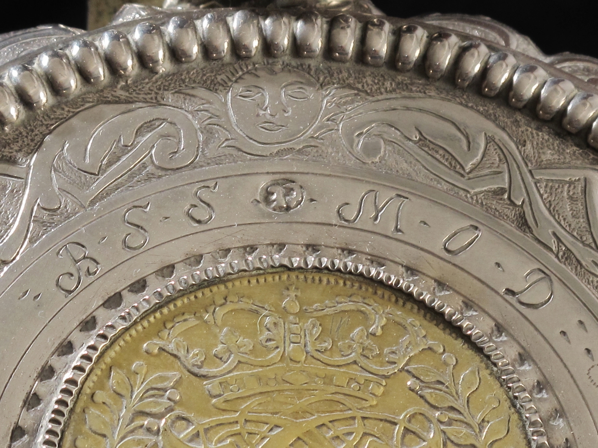 Stentøykrus m. sølvlokk. Lokket har en profilert, knekket kant m. regenceornamentikk, flat  toppflate, begrenset av opphøyet, tverriflet ring,  graverte blad og knekkete bånd på punslet bunn, i midten  mynt i smal riflet innf., som lokkgrep stående blad  m. opprullede fliker. Mynten C5´s kronede speilmonogram omgitt av kryssede bladgrener, reversen kronet riksvaaben og   IIII MARCK DANSKE 1694,  forgyllt. Rundt mynten gravert BSS, MOD, 1748, nyere innprikket FBW. Stpl. CE  i ovalt felt. 
Sølvlokket er montert på stentøykrus, som er sylindrisk m. svakt buede sider, lysbrun  glasur m. forsenket hvit ring øverst og nederst, samt hvit bunn,  munningsring og hank,  denne liten, flattrykkt øreformet.