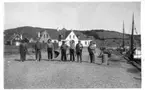 'Kattegattexpeditionen 1933: ::  :: ''Landexpedition till östsidan.'' 7 st män stående på rad framför hus vid en hamn. Bärande redskap. Enligt anteckning på fotografiets baksida: ''Fr. v. Skeppare Karl Johansson, mag. /... Elisason, båtsman Johan Johansson, Bror Johansson, agronom Gösta Hyle, Dr Harlad Alander, Leonard Axel Jägerskiöld. Länst till höger'' ::  :: Ingår i serie med fotonr. 2804-2826.'