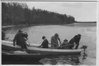 'Biologiska föreningens utfärd till Tåkern, Vadstena m.fl. platser: ::  :: Gruppen, 8 st hattklädda män,  på väg upp i båten. Gruppen har lagt till med 2 båtar med åror och ev. motor. Vy med sjön och skog. ::  :: Se fotonr. 3588-3634 samt 3661-3677.'
