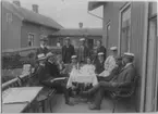 'Kristinebergs Zoologiska station. ::  :: Gruppfoto vid kaffebord på en veranda, 3 kvinnor, 10 män. Sittande på träbänkar, iklädda hatt, skärmmössa, studentmössa.I bakgrunden är ytterligare hus synliga samt ytterligare 2 personer. ::  :: Ingår i serie med fotonr. 5103:1-24.'