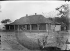 'Diverse fotografier från bl.a. dåvarande Nordrhodesia, nu Zambia, tagna av Konsul Magnus Leijer. ::  :: En tegelbyggnad fotograferad från avstånd. På verandan sitter 1 st kvinna och 1 st man och vid husknuten står 1 st  man. Även 2 hundar på verandan.'