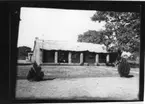 'Diverse fotografier från bl.a. dåvarande Nordrhodesia, nu Zambia, tagna av Konsul Magnus Leijer. ::  :: Tegelbyggnad med veranda fotograferad från avstånd, på verandan är en brittisk flagga synlig, bakom huset är 2 personer synliga.'