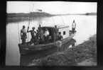 'Diverse fotografier från bl.a. dåvarande Nordrhodesia, nu Zambia, tagna av Konsul Magnus Leijer. ::  :: 8 st män på och invid en mindre båt med däckhus. 4 st av männen håller i pinnar, spjut(?), bland dem är 1 st kvinna stående.'