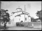 'Stor byggnad, kyrka med murad mur. Framför muren grupp människor, män, barn ev. vid en vattenbrunn. Telefonstolpe? ::  :: Ingår i serie med fotonr. 5275:1-21. Se även fotonr. 5269-5292 med bilder från Stuxbergs resa till Kaukasien.'