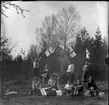 'Gruppbild med 11 flickor iklädda kjol. Håller flaggor. Picknick, matsäck. I färd med att äta. Glasflaskor. Smörgås. ::  :: Ingår i serie med fotonr. 5415:1-10.'