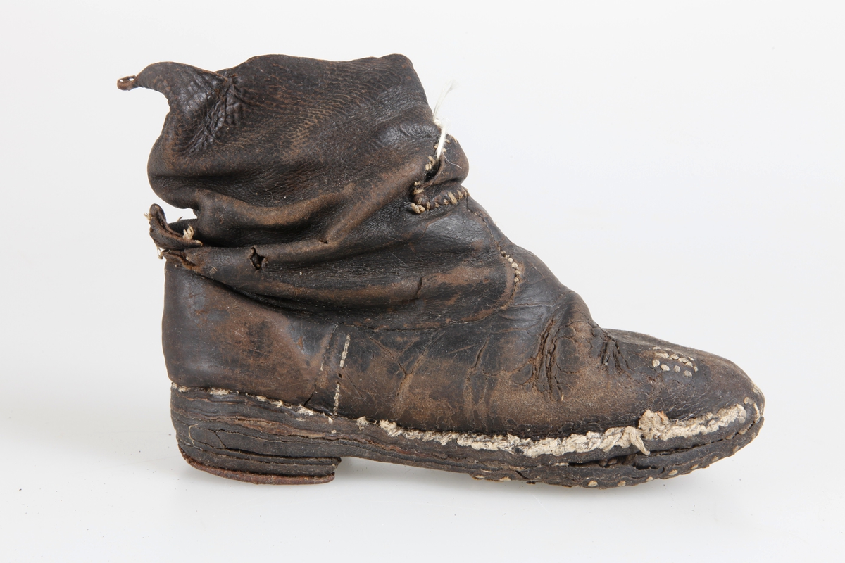 Udda barnkänga. Enläst. Brunsvart läder, nävermellansula. Handsydd. Randsydd, reseft, söm bak. Träpliggad sula och klack, klackjärn. Skaftet avskuret. Trasig, lagad.