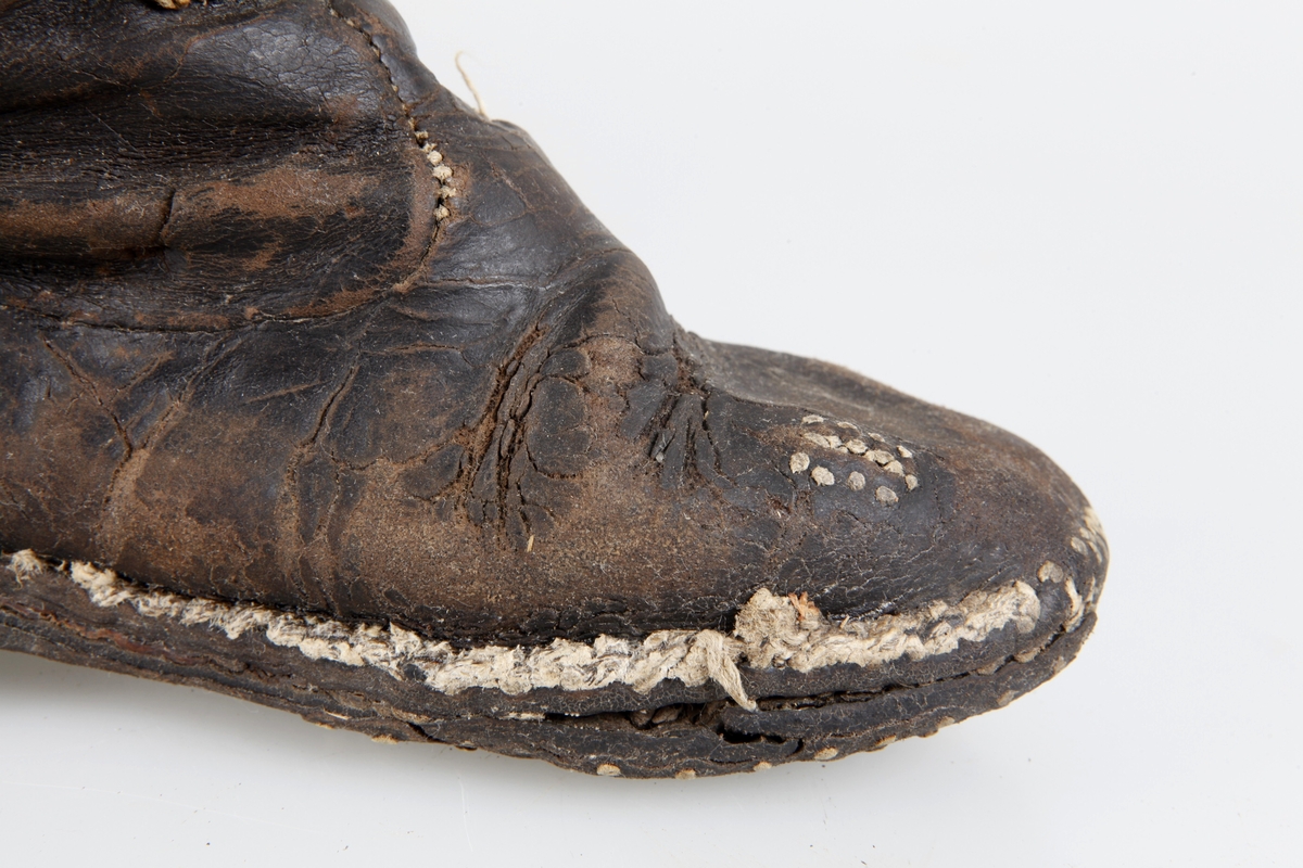 Udda barnkänga. Enläst. Brunsvart läder, nävermellansula. Handsydd. Randsydd, reseft, söm bak. Träpliggad sula och klack, klackjärn. Skaftet avskuret. Trasig, lagad.