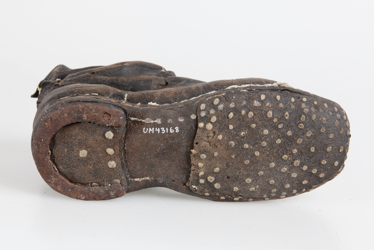 Udda barnkänga. Enläst. Brunsvart läder, nävermellansula. Handsydd. Randsydd, reseft, söm bak. Träpliggad sula och klack, klackjärn. Skaftet avskuret. Trasig, lagad.