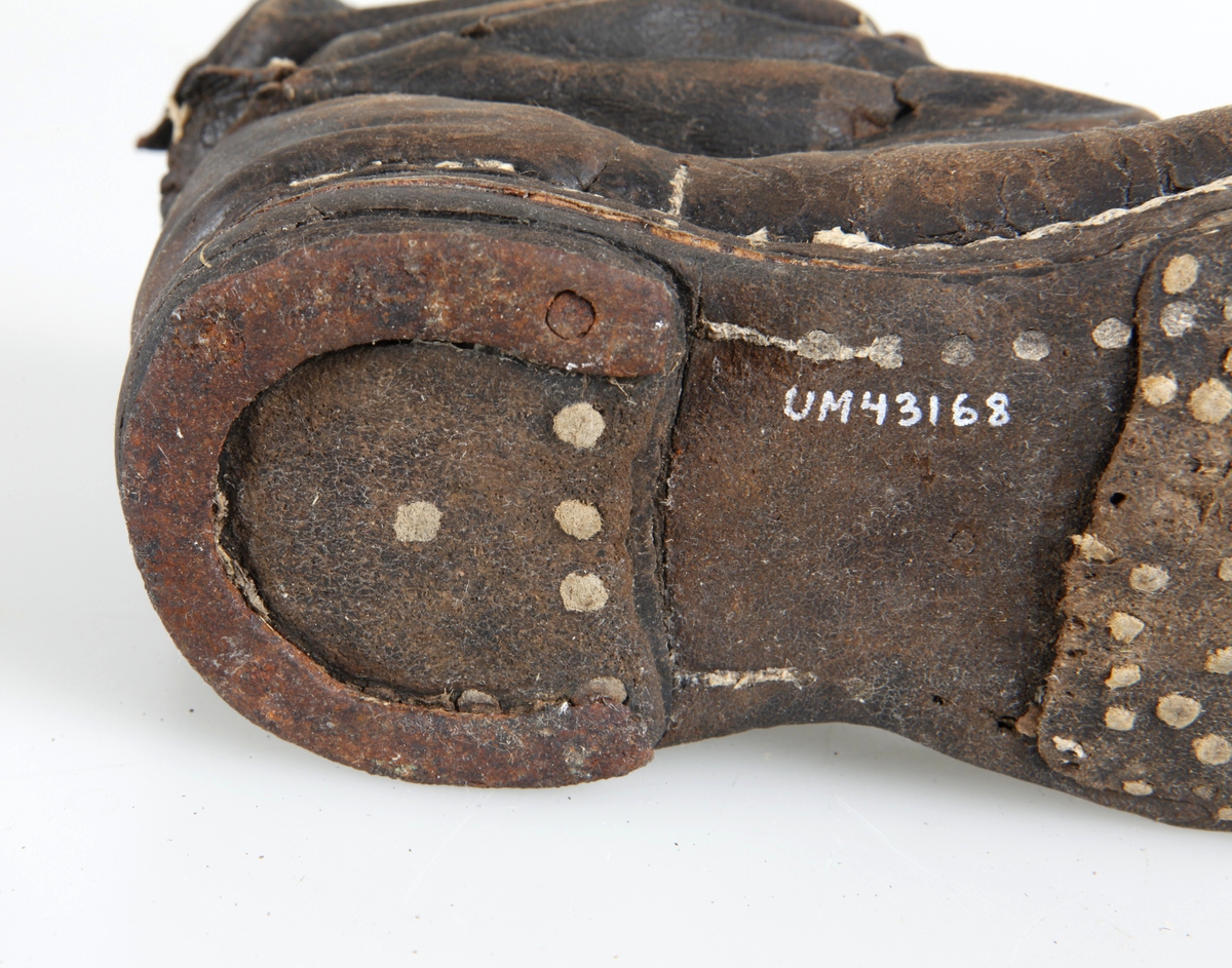 Udda barnkänga. Enläst. Brunsvart läder, nävermellansula. Handsydd. Randsydd, reseft, söm bak. Träpliggad sula och klack, klackjärn. Skaftet avskuret. Trasig, lagad.