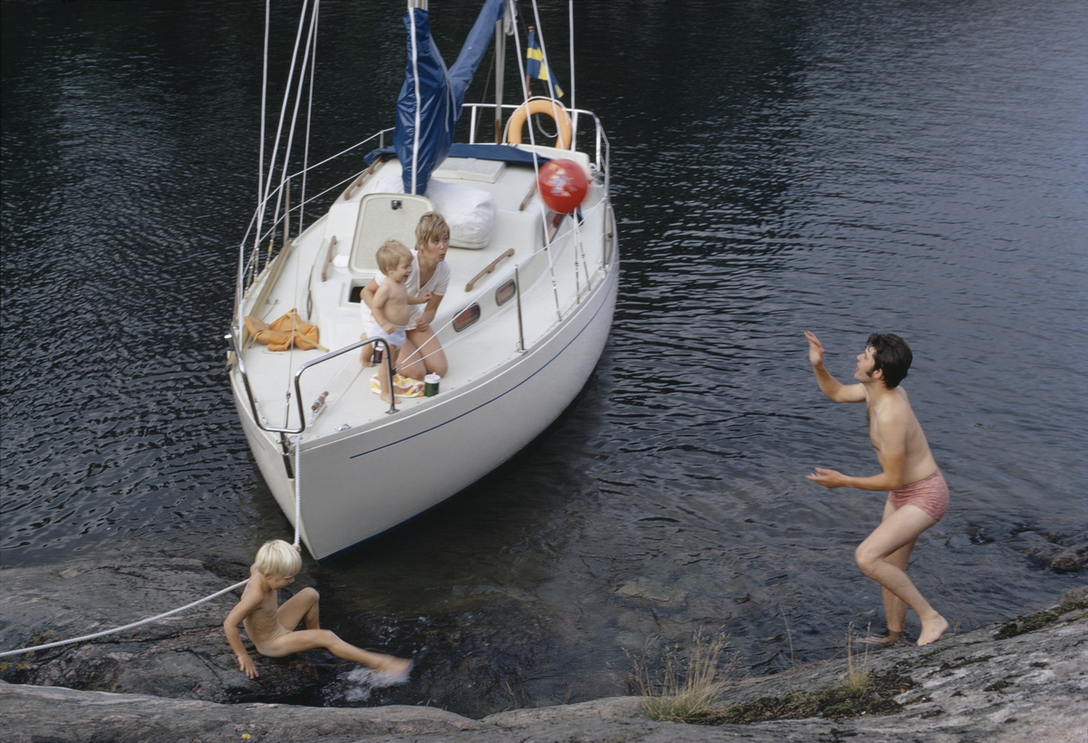 Albin Vega förtöjd vid klippa, barnfamilj leker med badboll. Marknadsföringbild.