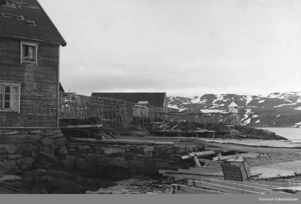 Båtsfjord etter krigen. I forgrunnen en ødelagt kai, i bakgrunnen tomme fiskehjeller og et fyr. Til venstre en bygning.