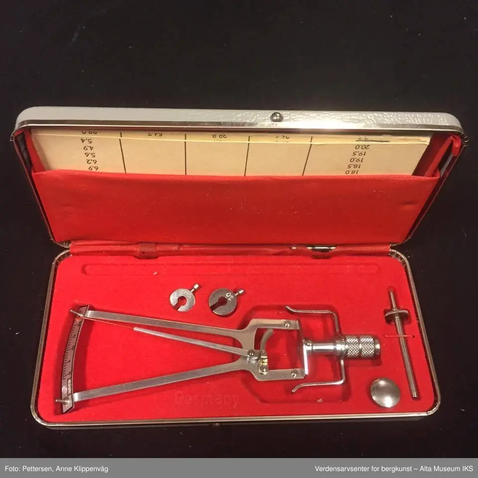 Rektangulær grå eske med rød foring. Inneholder et tonometer med tilleggsutstyr, en måletabell/bruksanvisning og et sertifikat.