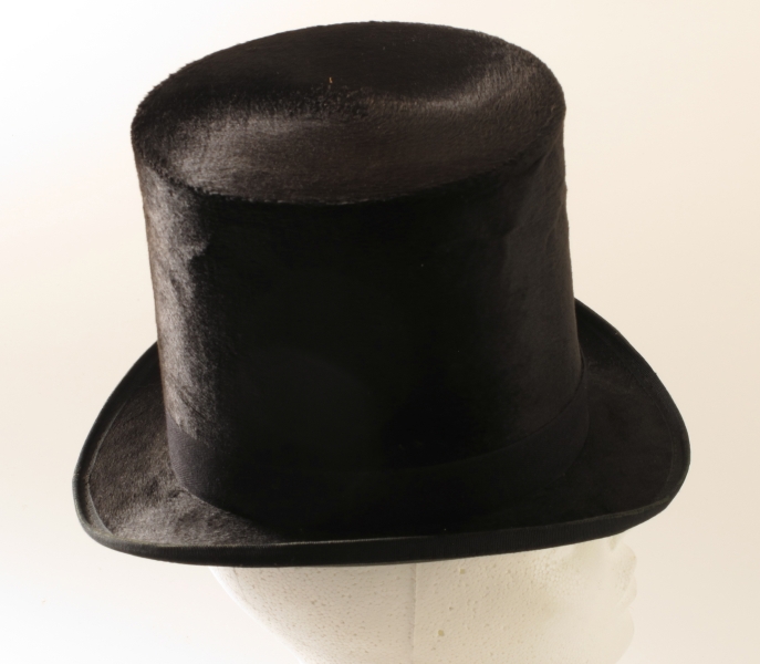 Flosshatt i hatteeske, merket hattemaker P. A. Paulsen, Arendal. a) Flosshatt. Sort floss, purpurfiolett og vinrødt for, trykt med gull midt i bunnen: P. A. Paulsen, Arendal. Svettebrem av konjakkfarget lær. Sort ripsbånd m. sløyfe.   
b) Hatteeske. Brunt lær, lærhank med messingbeslag, hvorpå innripet: T. Tellefsen. Foret med gammelrosa fløyel, innv. i bremmen gammelrosa tynt stoff.