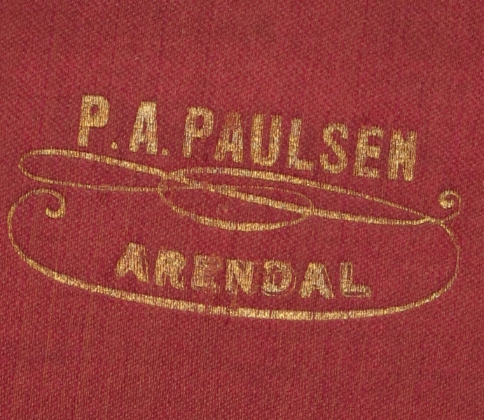 Flosshatt i hatteeske, merket hattemaker P. A. Paulsen, Arendal. a) Flosshatt. Sort floss, purpurfiolett og vinrødt for, trykt med gull midt i bunnen: P. A. Paulsen, Arendal. Svettebrem av konjakkfarget lær. Sort ripsbånd m. sløyfe.   
b) Hatteeske. Brunt lær, lærhank med messingbeslag, hvorpå innripet: T. Tellefsen. Foret med gammelrosa fløyel, innv. i bremmen gammelrosa tynt stoff.