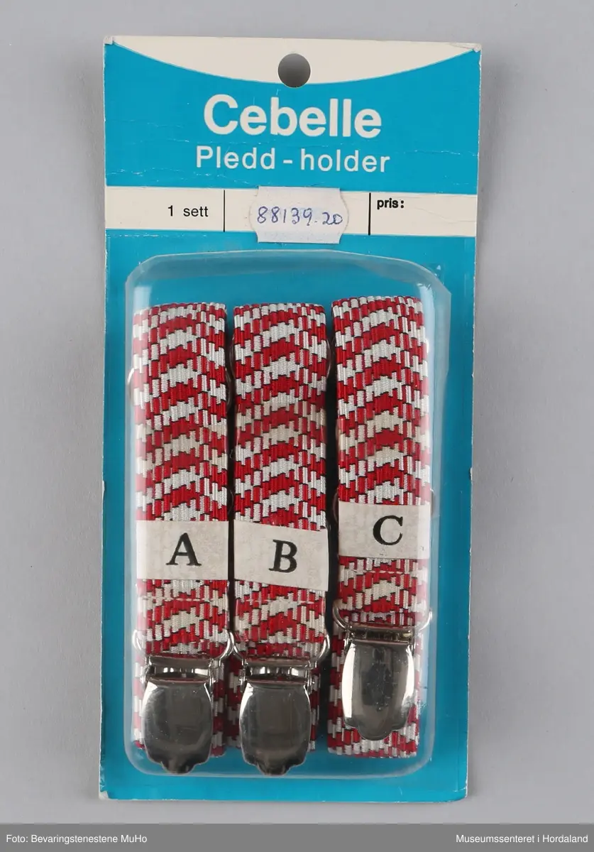 Eit sett pleddholdarar frå Cebelle, i uopna emballasje. Brukast til å feste pledd til t.d. ein sofa. Produsert av Oslo Baand & Lidsefabrik.