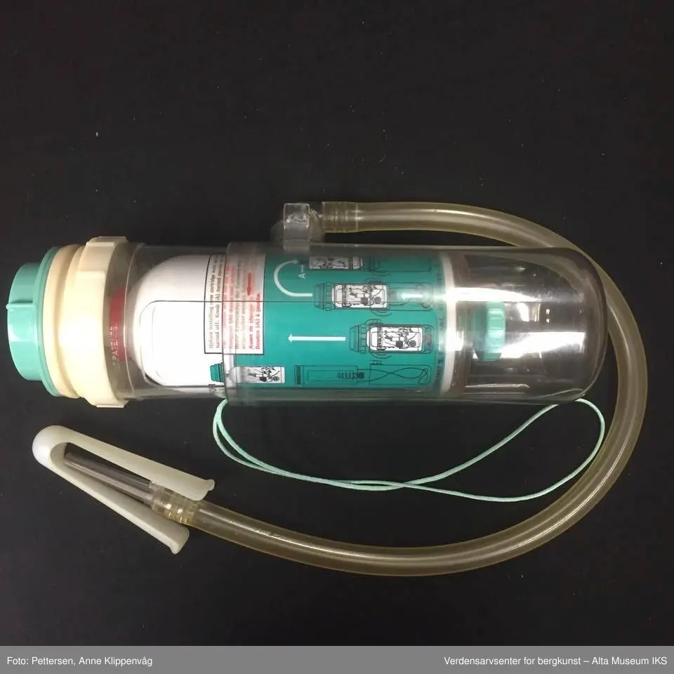 A. Sylinderformet plastapparat med tilfestet gummislange og gjennomsiktig plasttut.  Inneholder en driftspatron fylt med ufarlig gass.
B Rektangulært hefte, bruksanvisning.