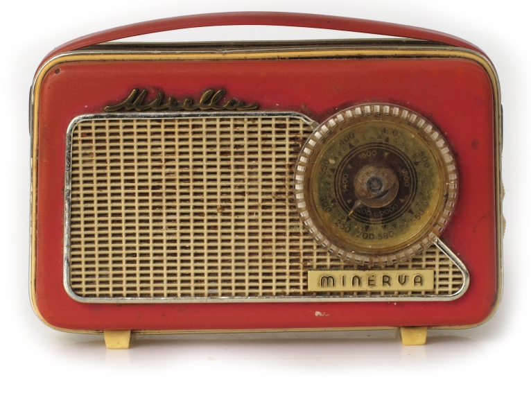 Liten reiseradio i originaleske. Smalt radioapparat  trukket med rosa og elfenbensfarget plast med bærestropp  av rosa plast lærimitasjon. Sirkelrund kanalsøker t. h. på ene langside, med høytalergitter t.v. Ligger i eske av elfenbenshvit kartong med gull , på "pute" trukket m. elfenbens filt.  Med garantikort fra Standard Radiofabrikk.