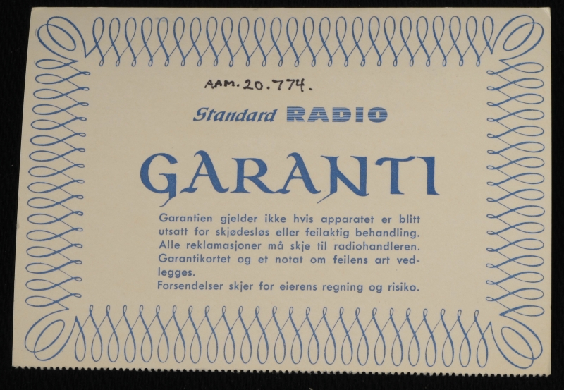 Liten reiseradio i originaleske. Smalt radioapparat  trukket med rosa og elfenbensfarget plast med bærestropp  av rosa plast lærimitasjon. Sirkelrund kanalsøker t. h. på ene langside, med høytalergitter t.v. Ligger i eske av elfenbenshvit kartong med gull , på "pute" trukket m. elfenbens filt.  Med garantikort fra Standard Radiofabrikk.