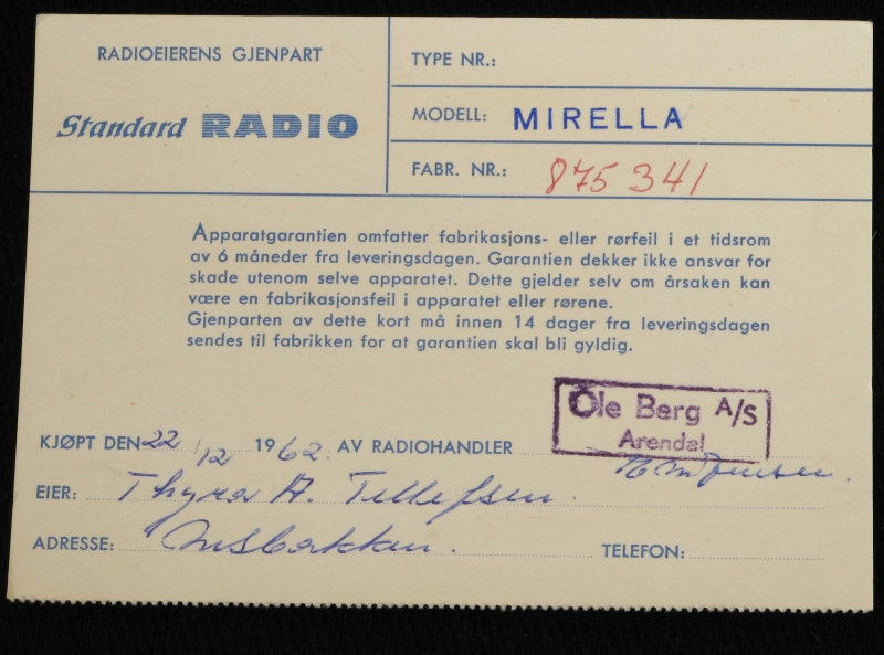 Liten reiseradio i originaleske. Smalt radioapparat  trukket med rosa og elfenbensfarget plast med bærestropp  av rosa plast lærimitasjon. Sirkelrund kanalsøker t. h. på ene langside, med høytalergitter t.v. Ligger i eske av elfenbenshvit kartong med gull , på "pute" trukket m. elfenbens filt.  Med garantikort fra Standard Radiofabrikk.