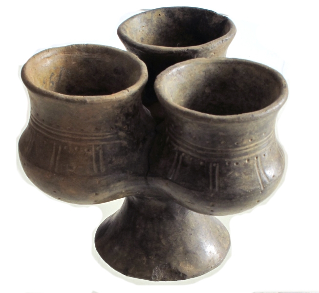 Tredelt fotbeger av brunlig til svart, forholdsvis tynt, godt brent gods med polert overflate, avbildet hos Bøe: anf. arb., fig. 186 (omtalt i teksten s. 130; men Bøe's datering er etter funnforholdene og de øvrige kar i sekundærgraven for tidlig (senere del av 4. årh.). Graven må dateres til 6. årh.). For de enkelte begrene som utgjør karet, har serien [Rygh fig.361]R. 361 stått som forbilde. Begrene har forholdsvis lang hals, markert bukknekk og ståflate. Der de støter sammen, er halsorneringen bøyet ned. Den består av en tredelt linjebunt på begge sider begrenset av en punktrekke. På skulderen er parvise vertikale linjebunter. På buken går de over i enkle punktrekker. Også ståflaten er avgrenset med samme ornering. Karene er kommuniserende. Sammenlimt. Høyde 9,9 cm, samlet tverrmål 11,2 cm. Begrenset høyde 5,9-6,2 cm, deres munningsdiam. 5,3-5,4 cm. (A. 196.)
