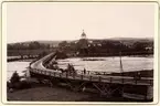 Skellefteå stad. Vy över kyrkan, älven och en träbro. På bron en häst och vagn.