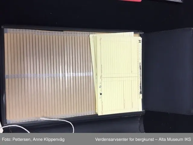 En rektangulær mappe i sort skinn som kan åpnes som en bok. På hver permside er det rammer i metall. I hver ramme er det 33 plastlommer med pappforing. På toppen av hver permside er det en lapp i sort skinn som skjuler den første plastlommen. Under skinnlappen på venstre permsiden er det liten bunke med gule arkivlapper som er håndskrevet på.
