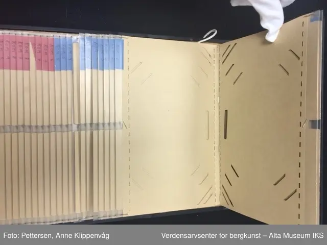 En rektangulær sort ramme i metall som er stiftet på et styrkke papp trukket i sort skinn. I rammen er det 49 plastlommer med pappforing. På toppen er det en lapp i sort skinn som skjuler den første plastlommen. På venstre siden av hver enkel plastlomme er det en liten farget lapp med påskrift.