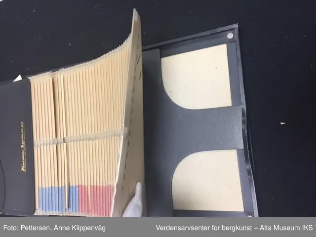En rektangulær sort ramme i metall som er stiftet på et styrkke papp trukket i sort skinn. I rammen er det 49 plastlommer med pappforing. På toppen er det en lapp i sort skinn som skjuler den første plastlommen. På venstre siden av hver enkel plastlomme er det en liten farget lapp med påskrift.
