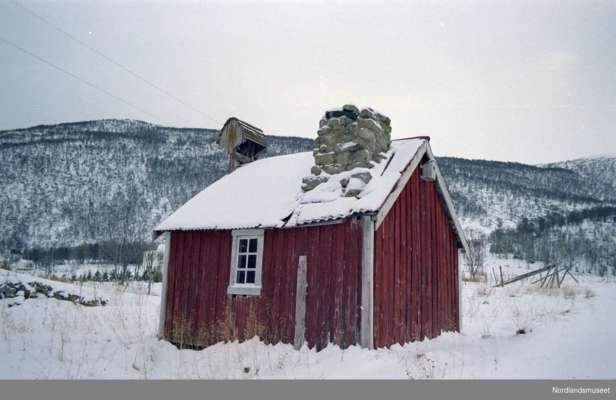 gammelt stabbur/eldhus. stående bordkledning, vinduer med småe vindusruter. fuglekasse på veggen. på taket stor pipe av stein og "stabbursklokke".