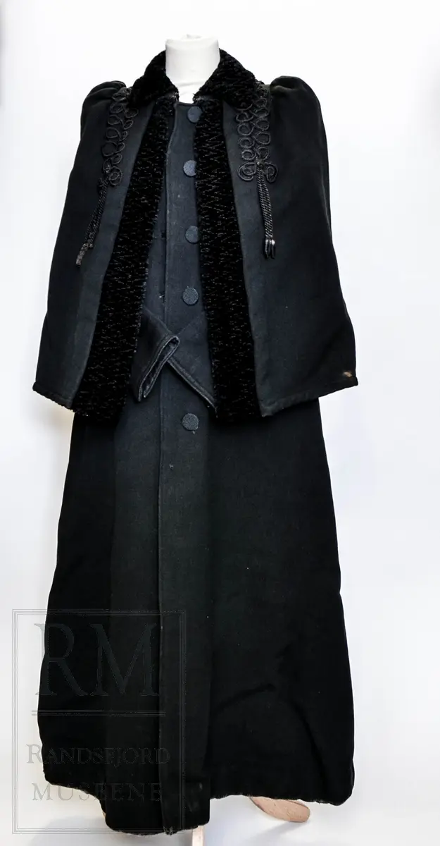 Sort kåpe i ullstoff med påsydd cape. Venstrekneppet, 6 stofftrukne knapper. Dekorert med flettede bånd (agraman). Lang, innsvinget modell med splitt bak. Uforet.
