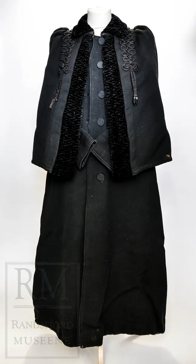Sort kåpe i ullstoff med påsydd cape. Venstrekneppet, 6 stofftrukne knapper. Dekorert med flettede bånd (agraman). Lang, innsvinget modell med splitt bak. Uforet.