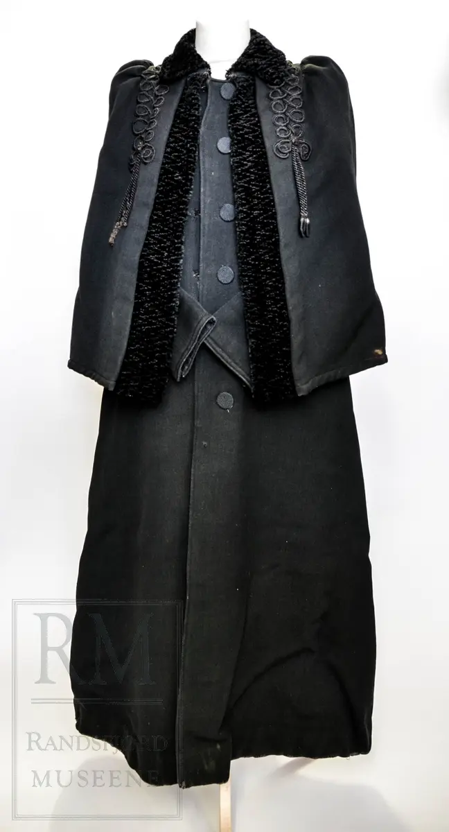 Sort kåpe i ullstoff med påsydd cape. Venstrekneppet, 6 stofftrukne knapper. Dekorert med flettede bånd (agraman). Lang, innsvinget modell med splitt bak. Uforet.