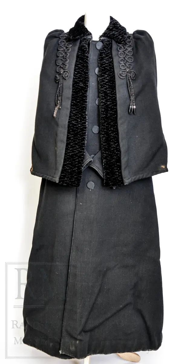 Sort kåpe i ullstoff med påsydd cape. Venstrekneppet, 6 stofftrukne knapper. Dekorert med flettede bånd (agraman). Lang, innsvinget modell med splitt bak. Uforet.
