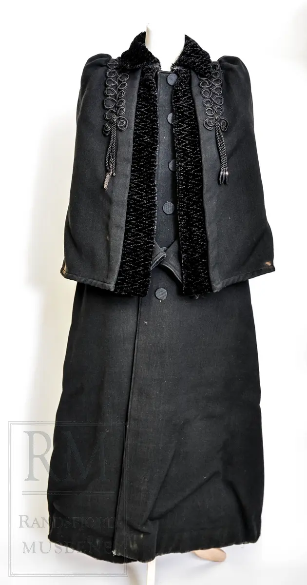 Sort kåpe i ullstoff med påsydd cape. Venstrekneppet, 6 stofftrukne knapper. Dekorert med flettede bånd (agraman). Lang, innsvinget modell med splitt bak. Uforet.