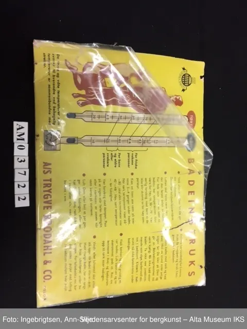 En rektangulær plakat i gul papp. Skrevet på med rød og svart skrift på høyre side. På venstre side er det en tegning med instruksjoner. Innpakket i plast.