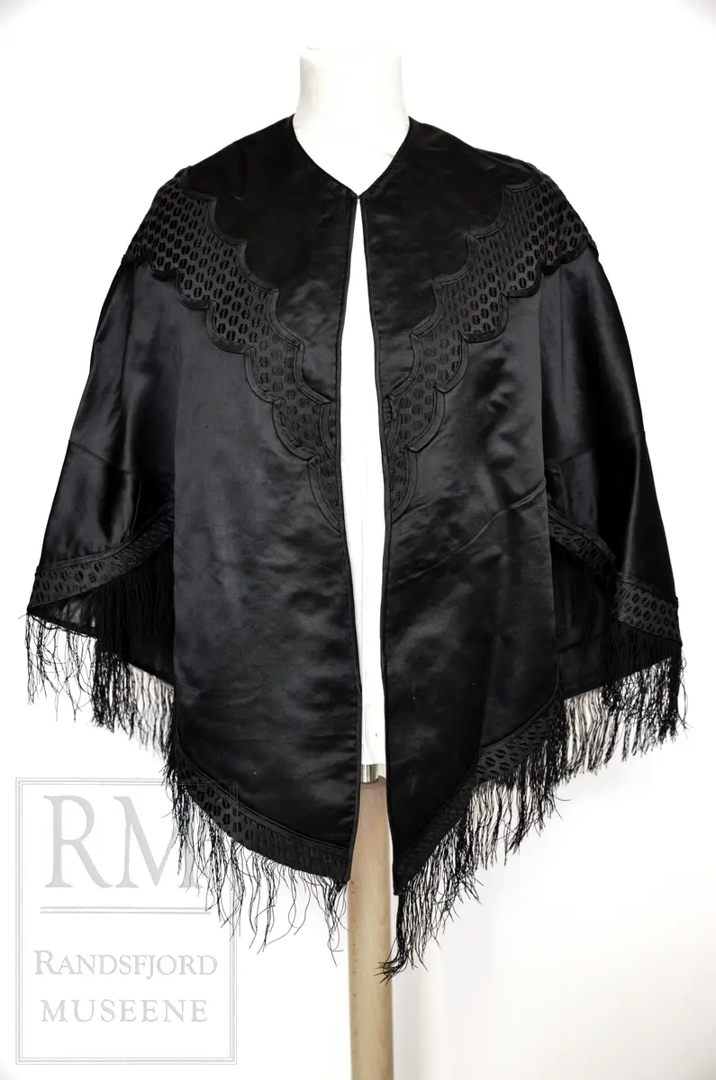 Håndsydd. Svart silkesatin med glanset bakside. Alpakkarenning (?) m / innslag i natursilke. Applikasjon av svart mønstret silkerips. To agramanrosetter med svarte glassperler. Sammensydd i rygg. Lukkes med hekte og malje ved halsåpning foran. Frynser.