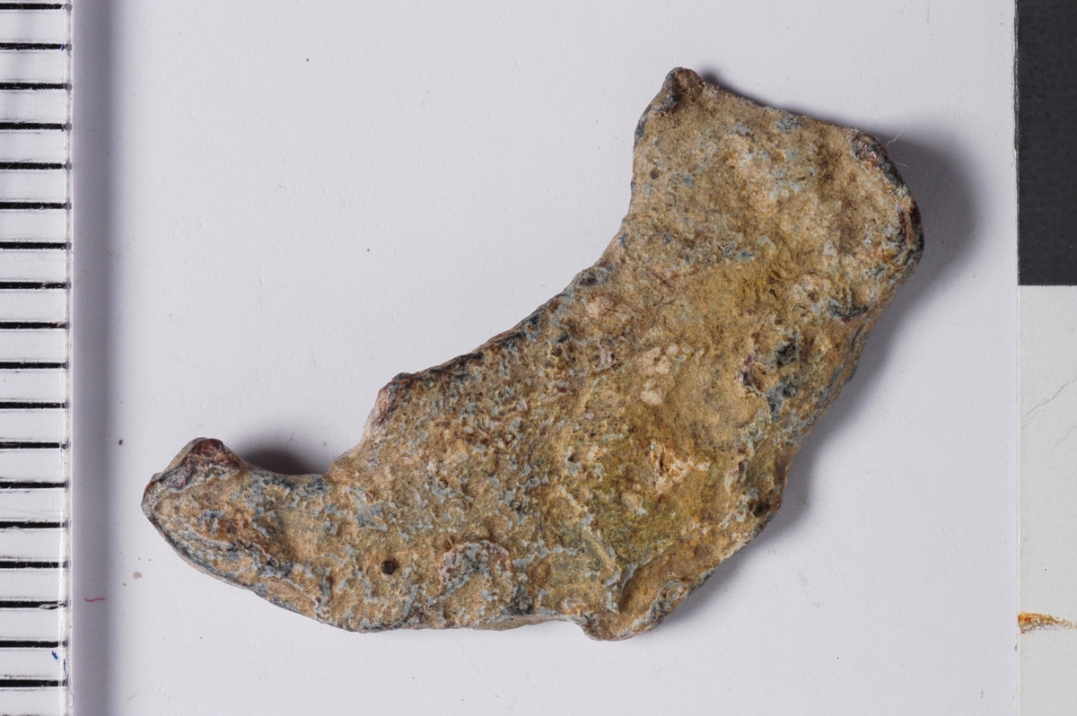 Del av plakett fra vareplombe, som har hatt to nagler. Gjenstanden har tegn til preg i form av perlerand i ytterkant. 