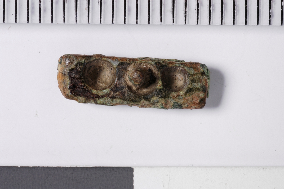 MALV: L. 12, 5 MM, 4 MM., BÅNDFORMET, SVAKT HVELVET PÅ EN SIDEN. 3 FORDYPNINGERHVOR DEN I MIDTEN HAR RESTER AV EN STIFT. 