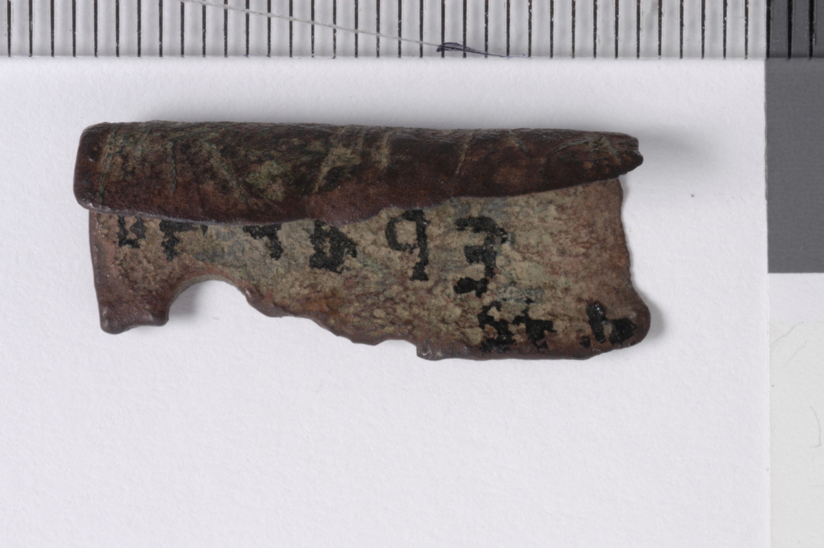 MALV: L. 25 MM, BÅNDFORMET FRAGMENT SOM ER BØYET. OVERSIDEN HARINNPRESSET DEKOR. DEN ENE ENDEN BEARBEIDET. 