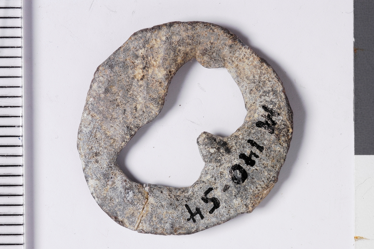 Del av vareplombe i form av en plakett med rest av streng. Gjenstanden har et hull i midten formet som et åttetall, noe som tilsier at vareplomben har hatt to nagler.  Det er tegn til preg samt en perlerad langs kanten av plaketten. Preget er utydet. 