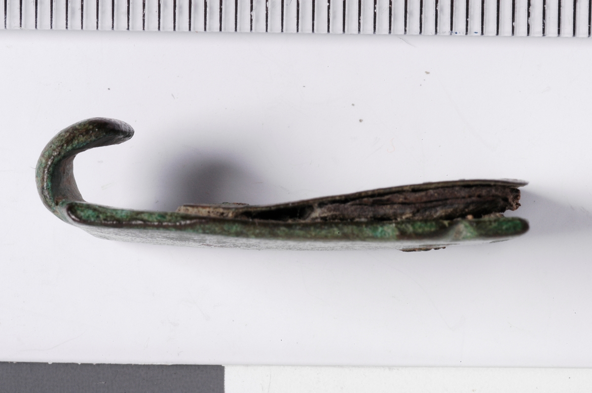 MALV: L. 28, 2  B. 18 MM, BANDFORMET MED HEMPE I ENE ENDEN. PÅ BAKSIDEN PÅSATTEN TYNN PLATE MED NAGLER. RESTER AV PERMEN SITTER IGJEN MELLOM SPENNEOG PLATEN, UTEN DEKOR. 