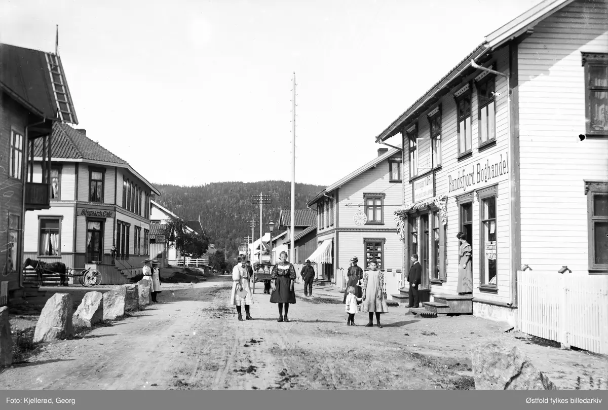 Gateparti fra Jevnaker. 
Skilt med tekst: 
Randsfjord Boghandel.
Uhrmager N. O. Rud (?)
Ødegaards kafe.
