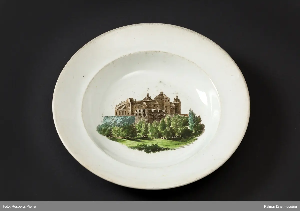 KLM 45642 Tallrik, av porslin. Djup tallrik med tämligen brett brätte. På undersidan text: Kalmar slott, samt tillverkarstämpel som inte kunnat tydas. Tryckt dekor med motiv av Kalmar slott i brunt och grönt, slottet inbäddat i grönska, nedanför vallarna buskar och träd. Motivet dateras till 1860-70-tal, eftersom tornhuven på Kuretornet omgestaltades ca 1880. Om motivet och tallriken är från samma tid, kan tallriken dateras till 1860-70-tal.