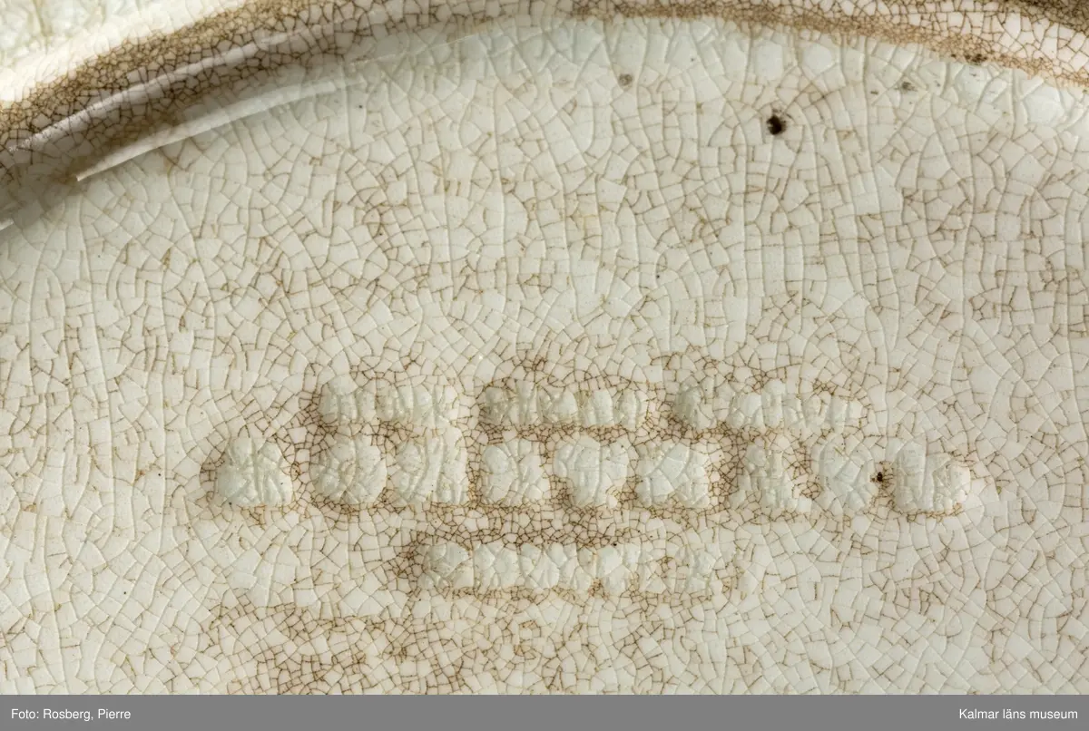KLM 45642 Tallrik, av porslin. Djup tallrik med tämligen brett brätte. På undersidan text: Kalmar slott, samt tillverkarstämpel som inte kunnat tydas. Tryckt dekor med motiv av Kalmar slott i brunt och grönt, slottet inbäddat i grönska, nedanför vallarna buskar och träd. Motivet dateras till 1860-70-tal, eftersom tornhuven på Kuretornet omgestaltades ca 1880. Om motivet och tallriken är från samma tid, kan tallriken dateras till 1860-70-tal.