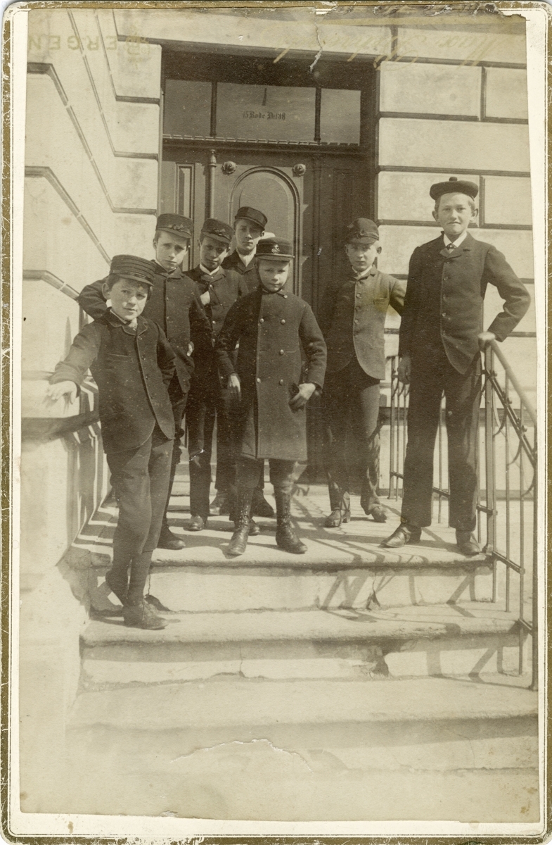 Sju gutar i trapp utanfor bygning i Langes gate på Sydneshaugen i Bergen i 1887. Emil Clausen heilt til høgre.