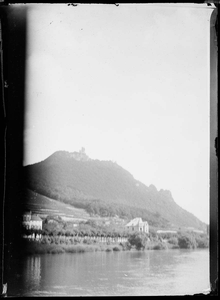 "Langenau 1, Luxemburg 2" (inskr eske). En åsside med et bygg på toppen og to hus ved vannet.