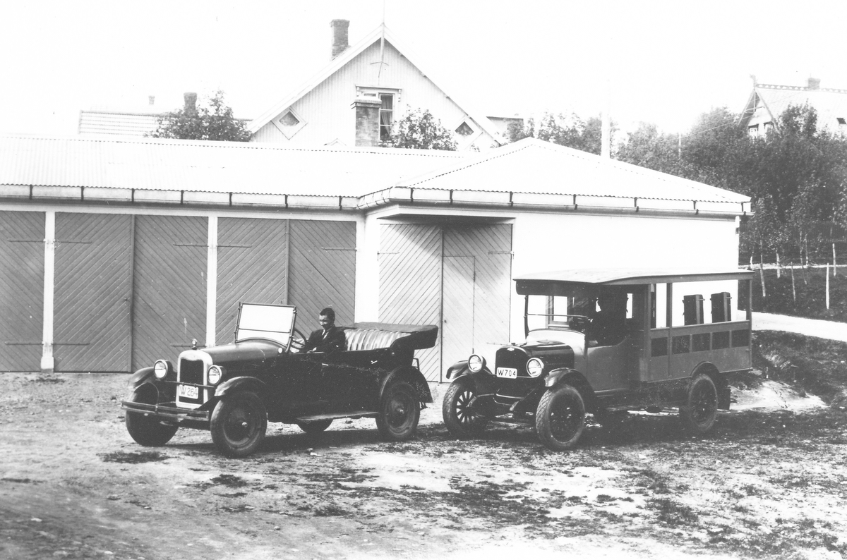 To biler foran garasjerekke. To Chevrolet. 1926 personbil registreringsnummer W-264 og 1928 buss W-704. Eier av begge: Håkon Eriksen Mo, iflg Bilboken for Norge 1930. Mann sitter i den ene bilen. Taket på ett hus i bakgrunnen.