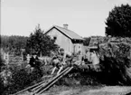 Träbearbetning (snickeri), en man som sågar, en man med dragspel, tre pojkar.
Bostadshus i bakgrunden.