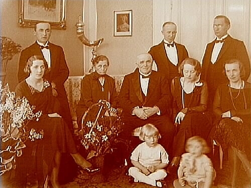 Rumsinteriör, grupp 10 personer.
Fabrikör Israel Eriksson. Villa Haganäs. 
Personerna på bilden är Israel Eriksson (1862-1933) och hans barn med respektive och barnbarn.
Till vänster står yngsta sonen Gustav Eriksson med hustru Margit.
I soffan sitter hembiträdet Emilie Larsson. Bredvid Emilie sitter Israel Eriksson.
Bakom Israel står hans son Oskar Eriksson och framför Oskar sitter hans hustru Margit.
Längst till höger sitter Israels dotter Greta Joelsson och bakom henne står hennes man Emil.
På golvet, till vänster, sitter Torsten Joelsson, son till Emil och Greta Joelsson.
Till höger sitter Britt-Louise Ursing (Eriksson), dotter till Oskar och Margit Eriksson.